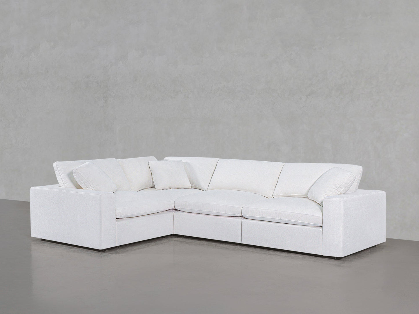 A 4-Seat Modular Corner Sectional with plush cushions and pillows sits on a polished concrete floor against a light gray textured wall.