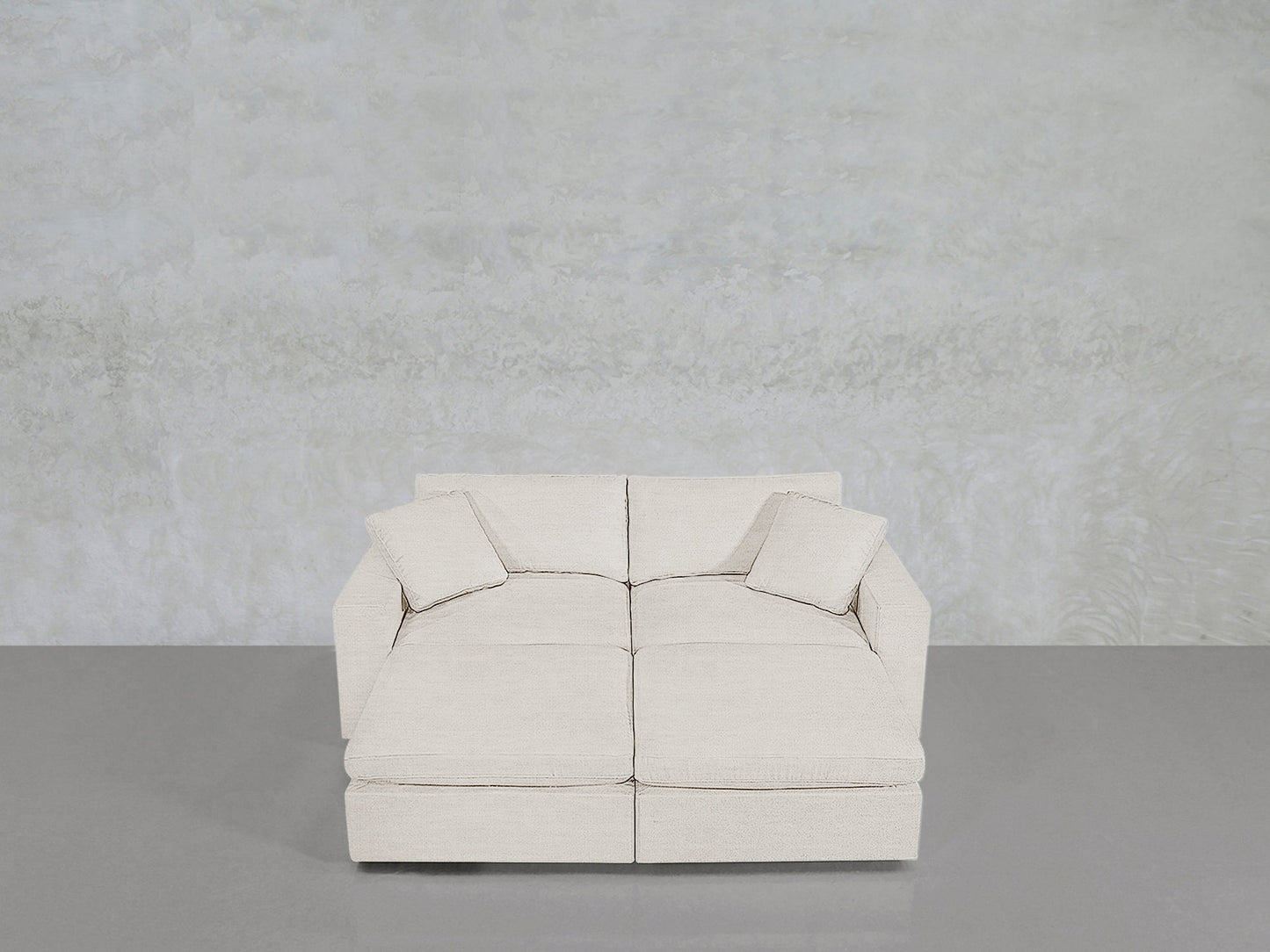A modern, light beige 4-Seat Modular Daybed with two matching square cushions, set against a textured light gray wall and a smooth gray floor.