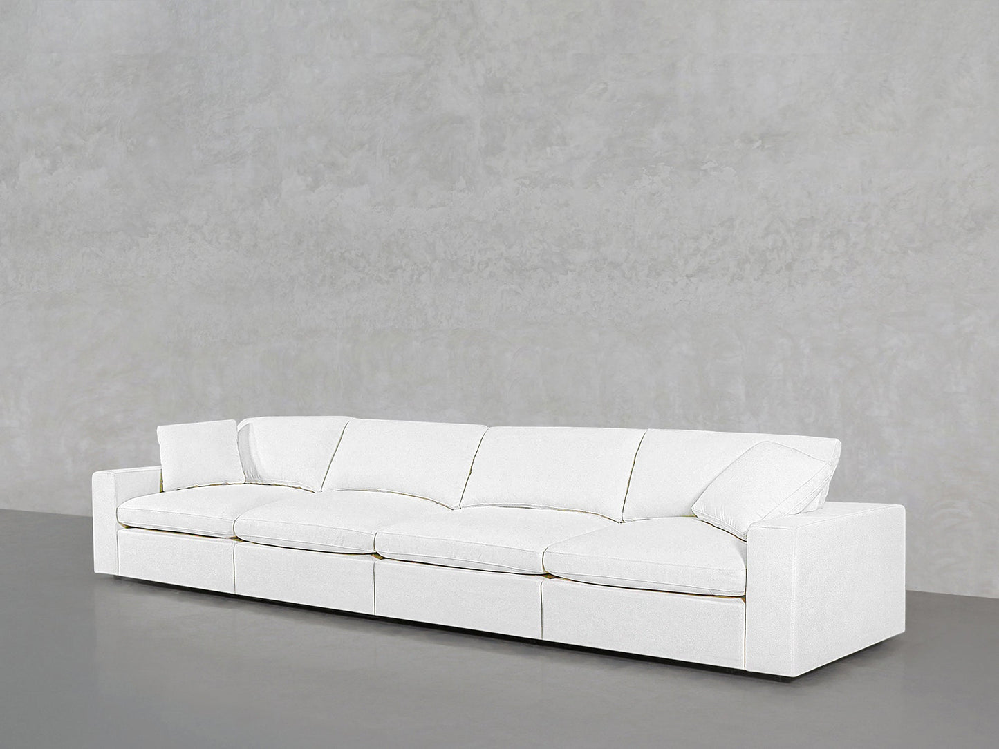 The 4-Seat Modular Sofa, featuring a modern and minimalist white design with four seat and back cushions, is placed against a gray concrete wall in an empty room.