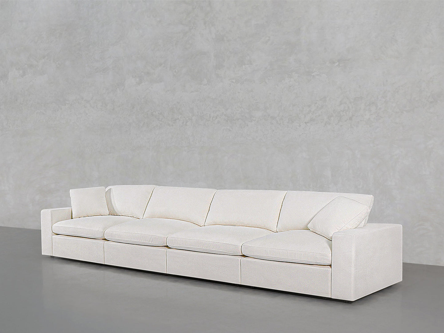 The 4-Seat Modular Sofa, featuring a modern white design with four seat cushions and two angled armrests, is set against a light gray textured wall on a gray floor.