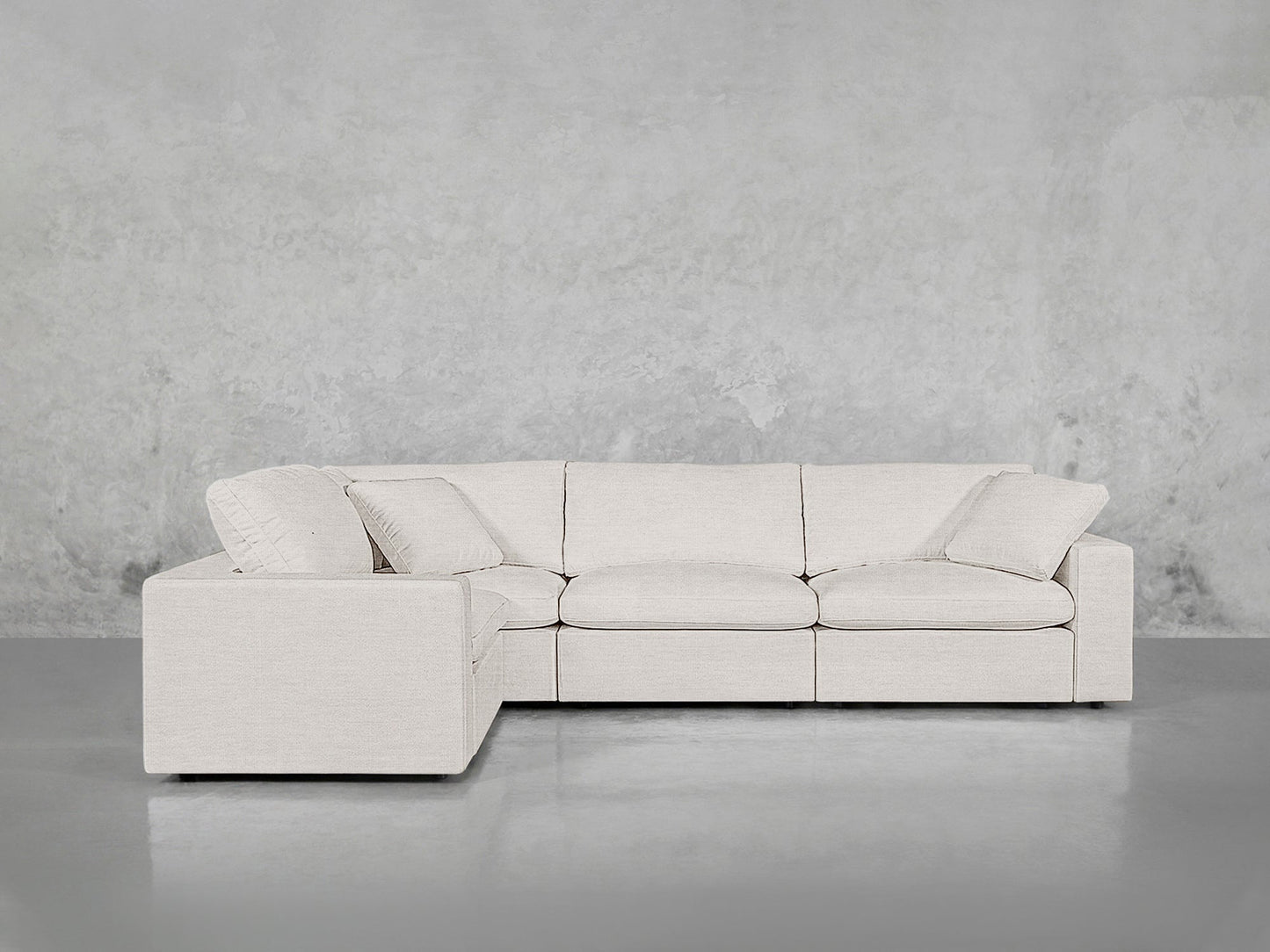 A modern 4-Seat Modular Corner Sectional in light beige with matching pillows is placed against a plain, light gray concrete wall and a reflective floor.