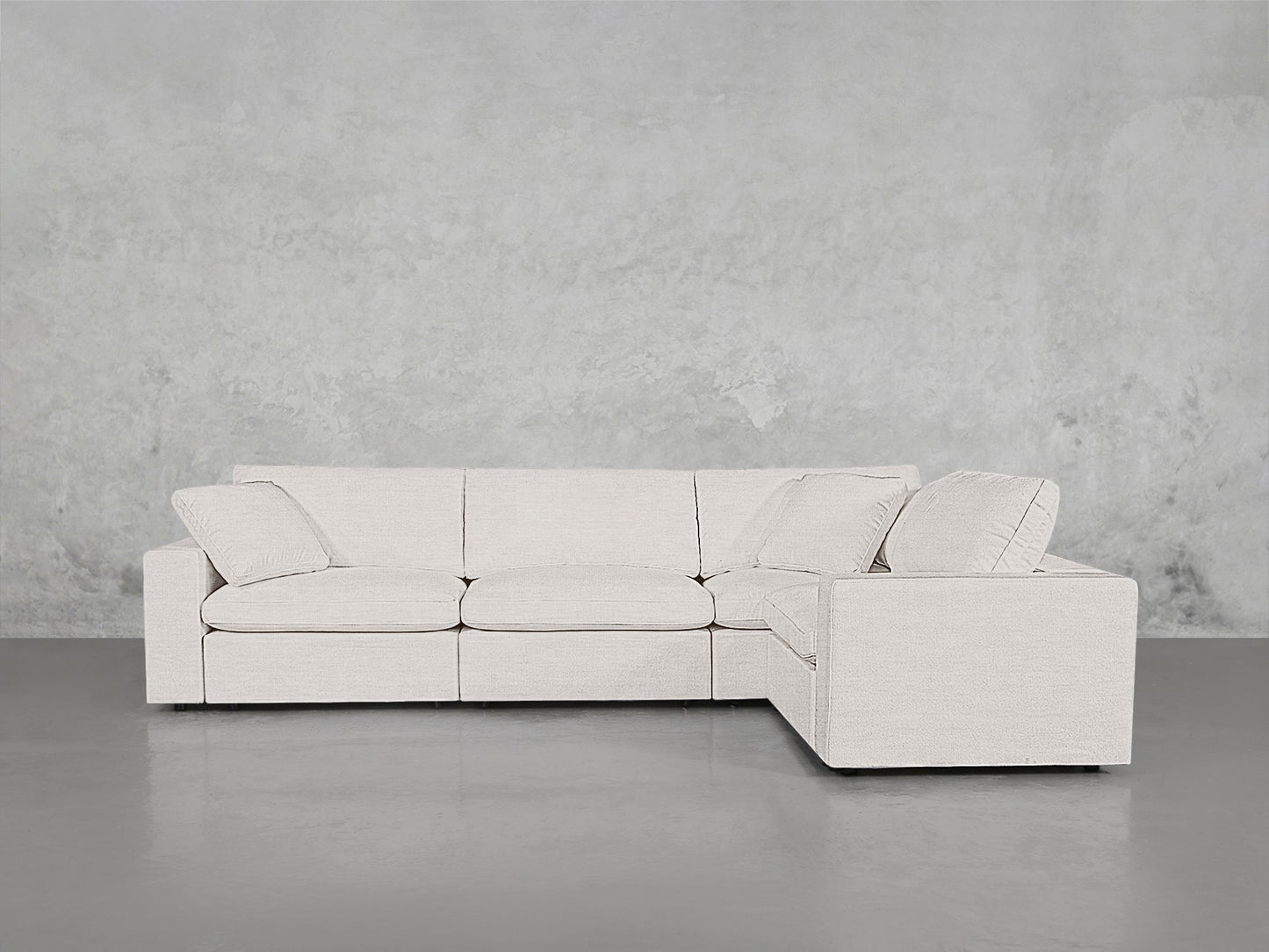The 4-Seat Modular Corner Sectional in light beige features clean lines and three seat cushions, set against a plain, textured gray wall and gray floor.