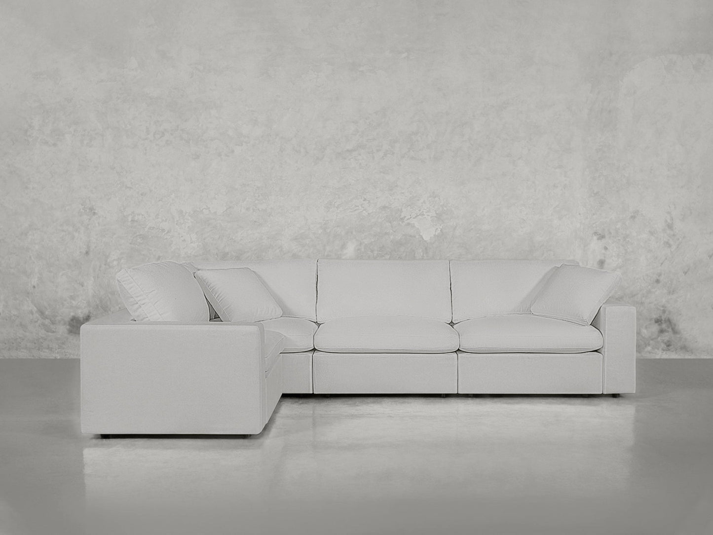 A 4-Seat Modular Corner Sectional in light gray, featuring three seat and back cushions, sits against a textured pale gray wall on a polished concrete floor.