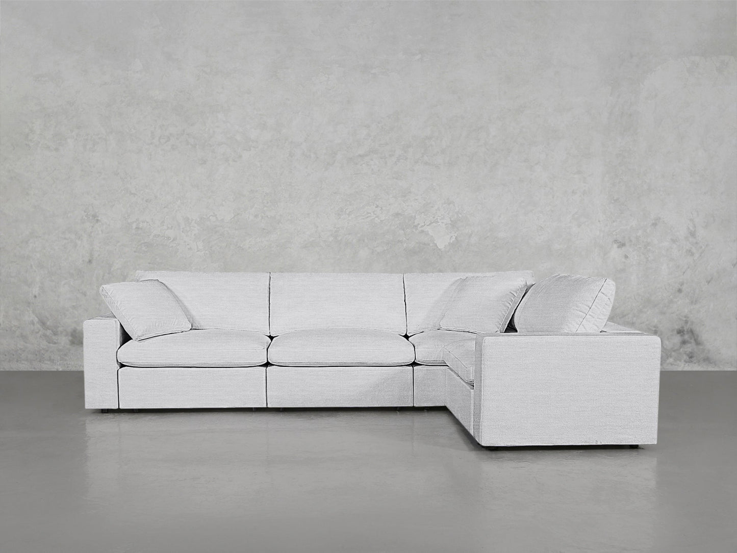 The 4-Seat Modular Corner Sectional, featuring three seat cushions, wide armrests, and back and side pillows for comfort, is shown in light gray on a smooth gray floor against a textured gray wall.