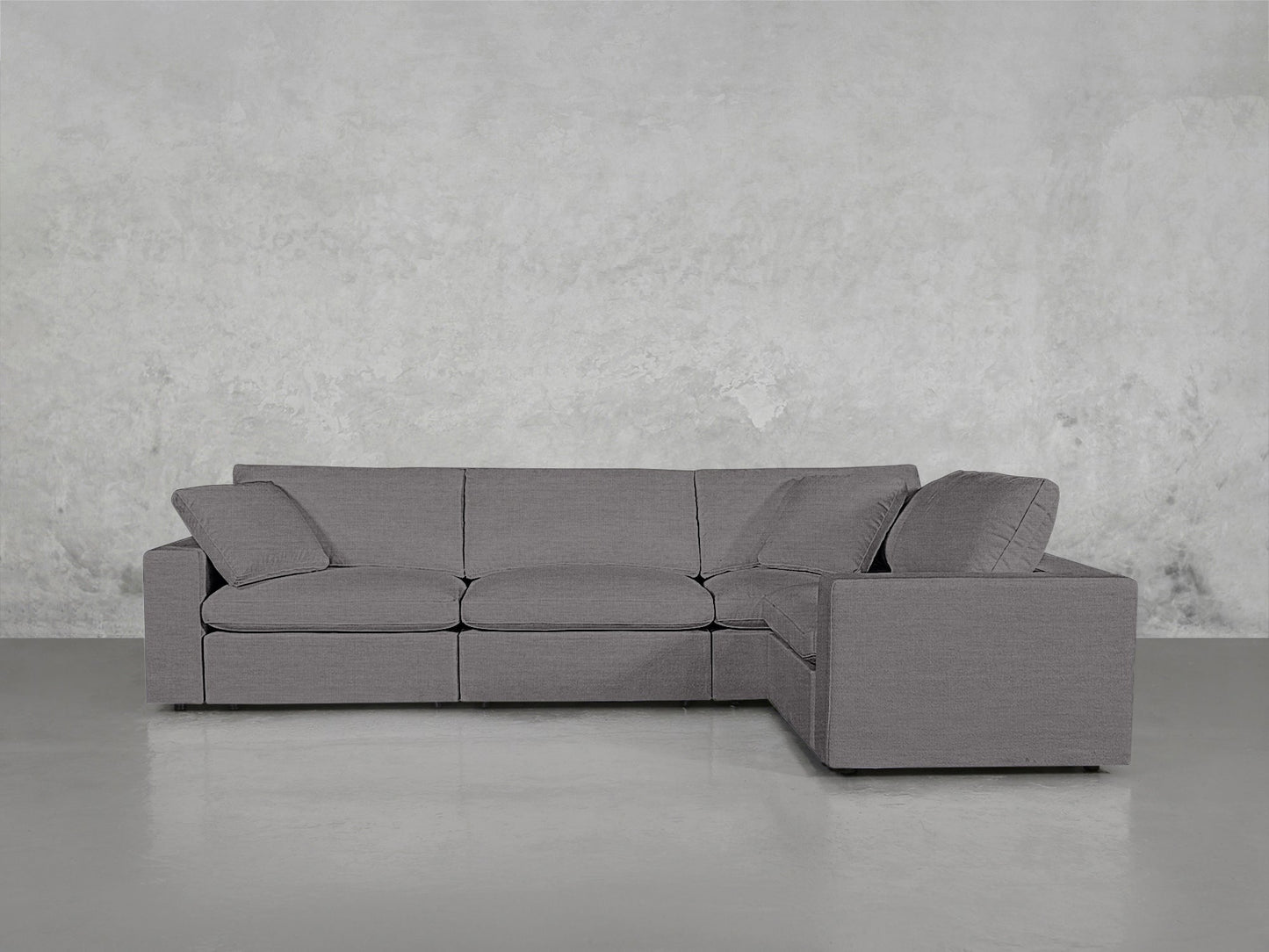 The 4-Seat Modular Corner Sectional, featuring clean lines and plush cushions in modern gray, is placed against a light gray textured wall and a smooth, light-colored floor.