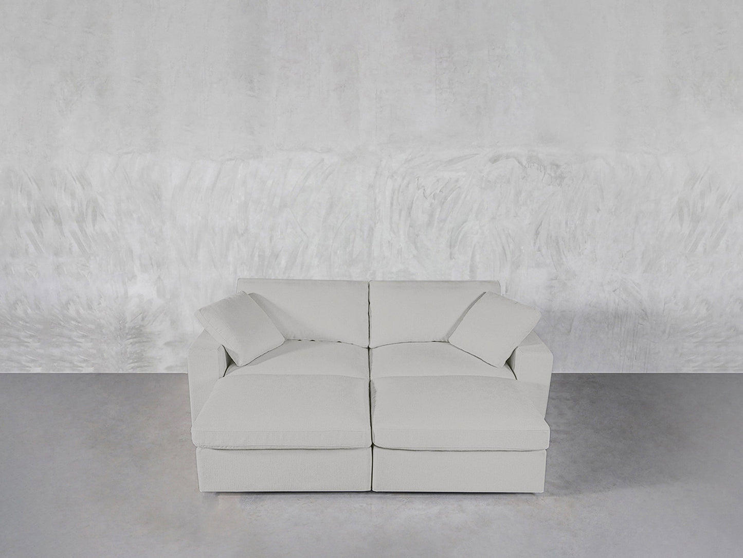 A modern light gray 4-Seat Modular Daybed with two seat cushions and two matching pillows sits against a textured light gray wall on a smooth gray floor.