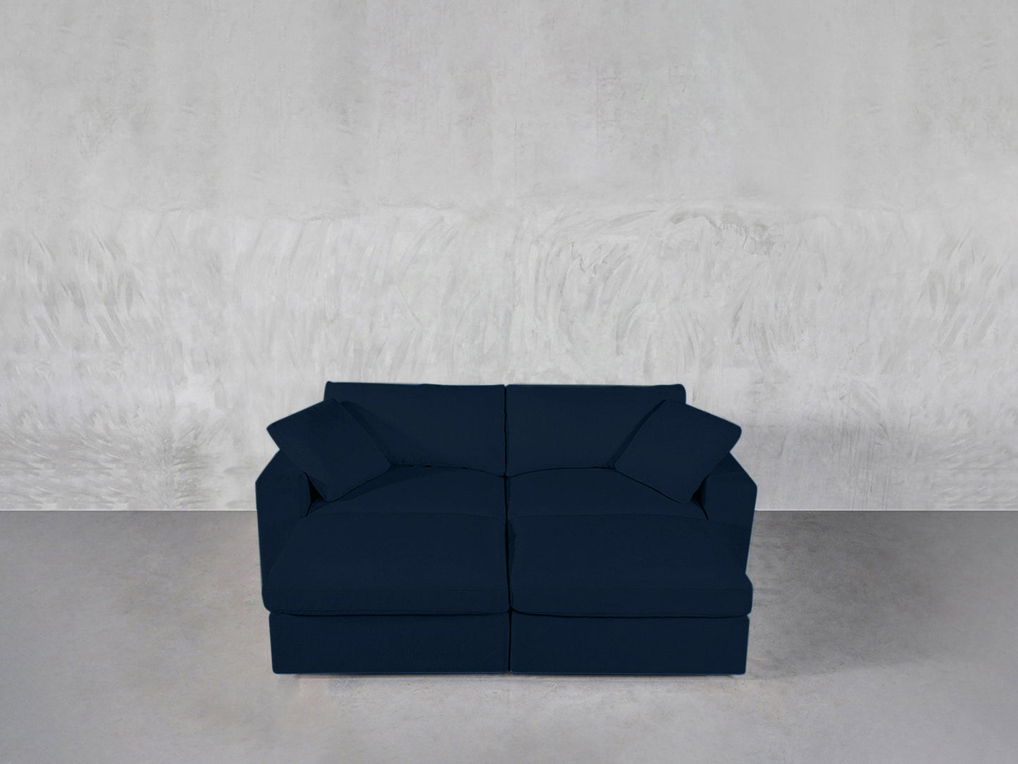 A 4-Seat Modular Daybed upholstered in dark blue with two matching cushions sits against a light gray textured wall on a smooth, neutral floor.