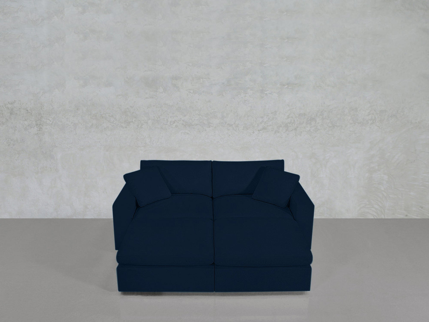 A 4-Seat Modular Daybed in dark blue with two matching square cushions sits against a light gray textured wall on a smooth, light-colored floor.