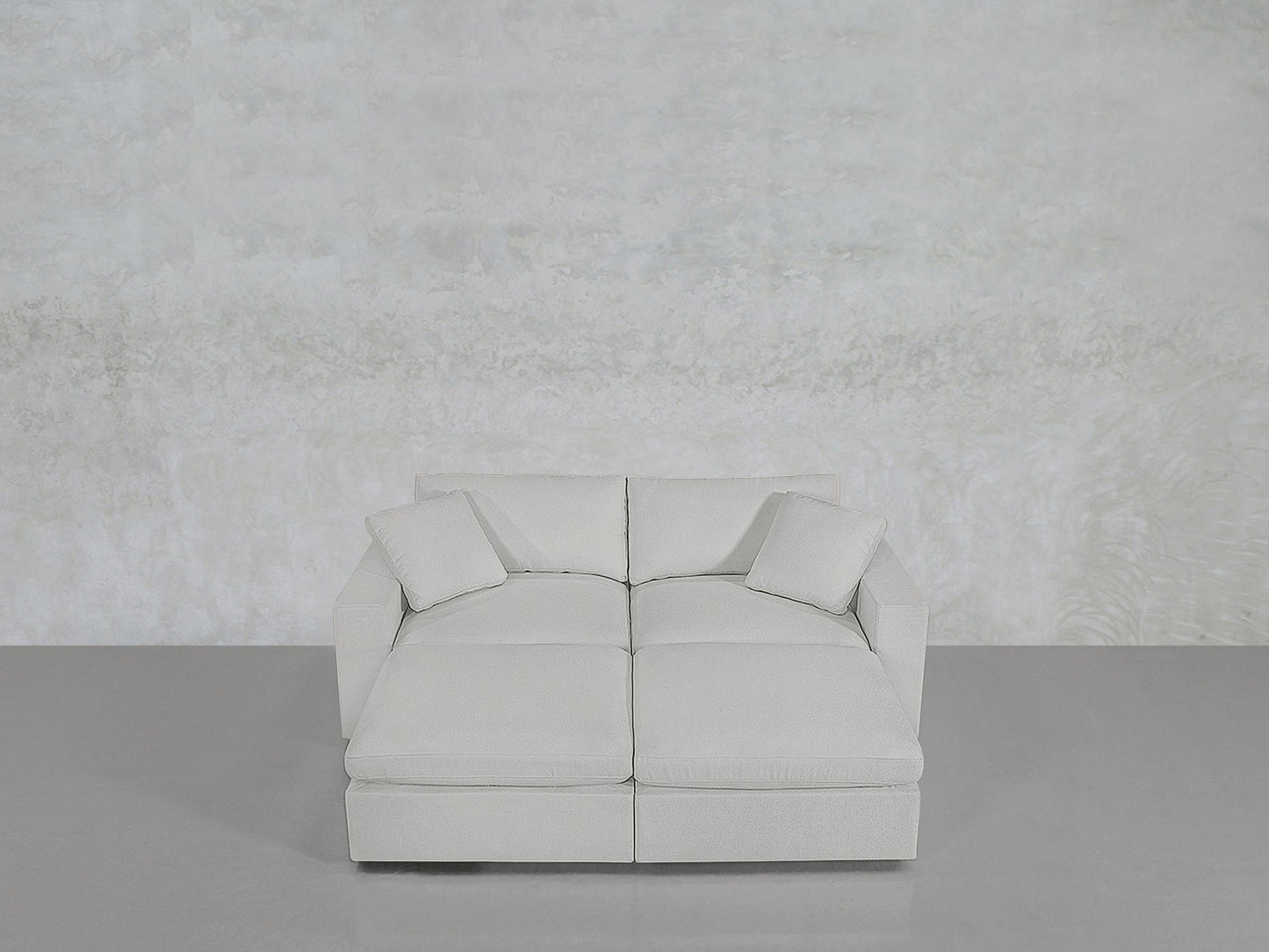 A 4-Seat Modular Daybed in light gray with two matching cushions is placed against a textured pale wall and a smooth gray floor.