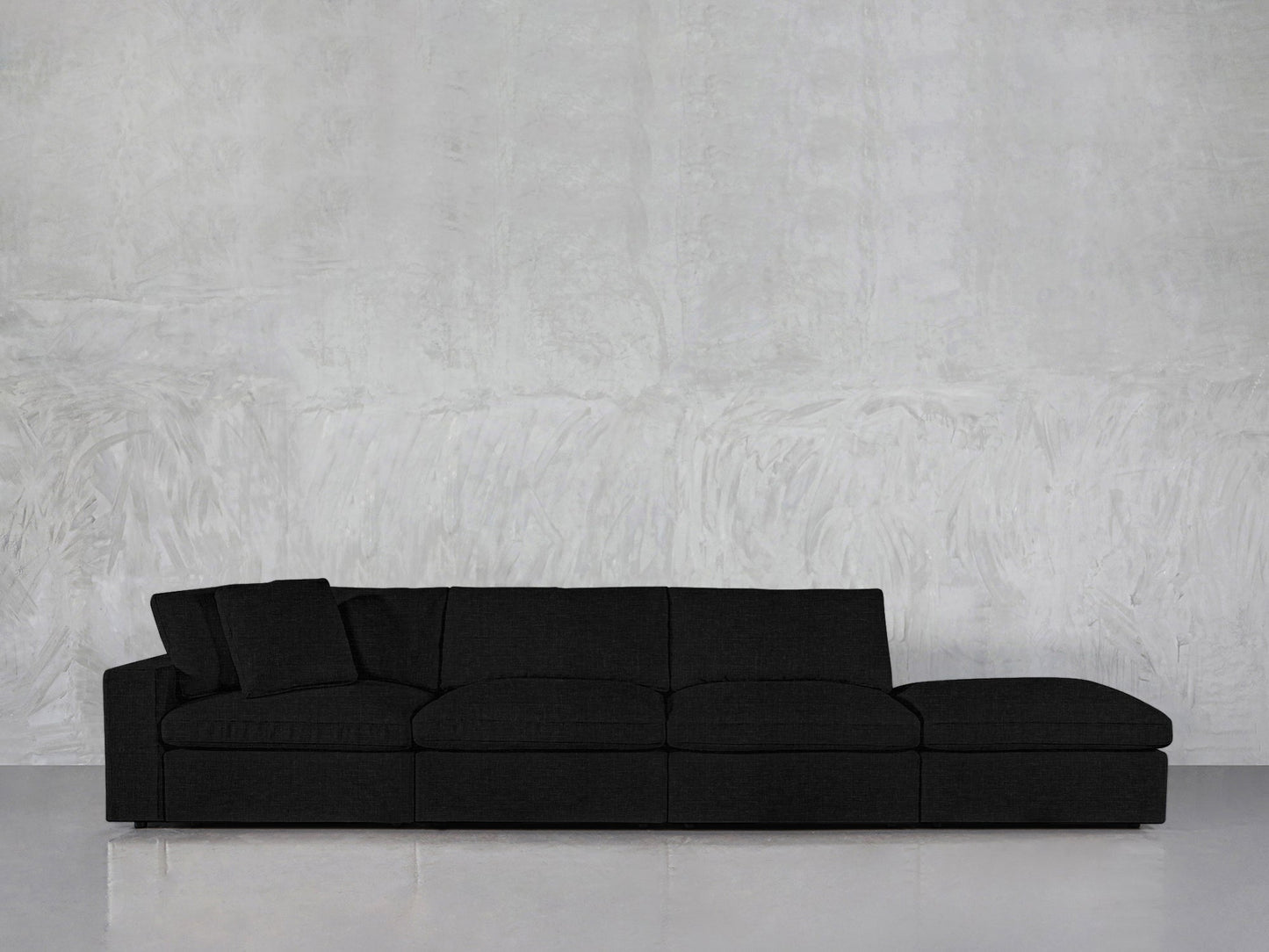 The 4-Seat Modular Lounger Sofa in dark gray features four seat sections and a matching throw pillow, displayed against a light gray textured wall and a light floor.