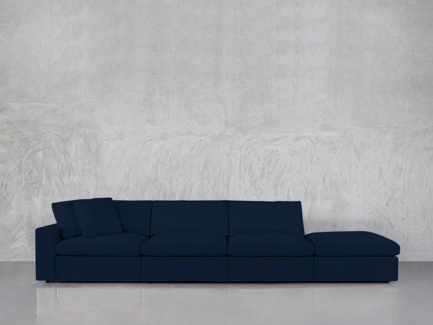 A modern, dark blue 4-Seat Modular Lounger Sofa with two back pillows is placed against a light gray textured wall on a reflective floor.