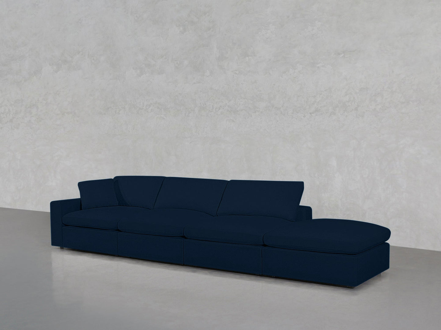 The 4-Seat Modular Lounger Sofa in dark blue, featuring wide seats and a chaise lounge, is placed against a light gray textured wall on a polished concrete floor.