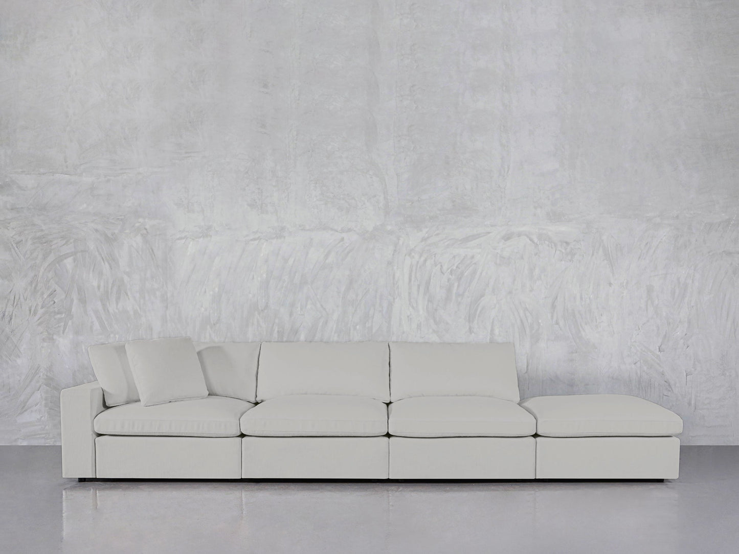 The 4-Seat Modular Lounger Sofa, in light gray with three seat cushions, a right-side chaise lounge, and matching pillows, sits against a textured gray wall on a shiny floor.