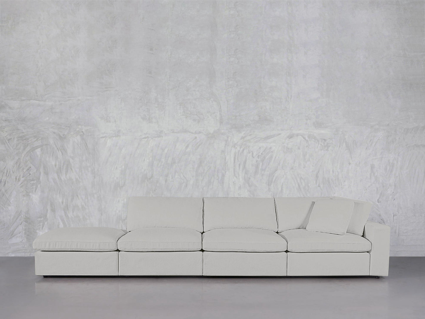 The 4-Seat Modular Lounger Sofa, in light gray with one right-side cushion, stands against a textured light gray wall and smooth gray flooring.