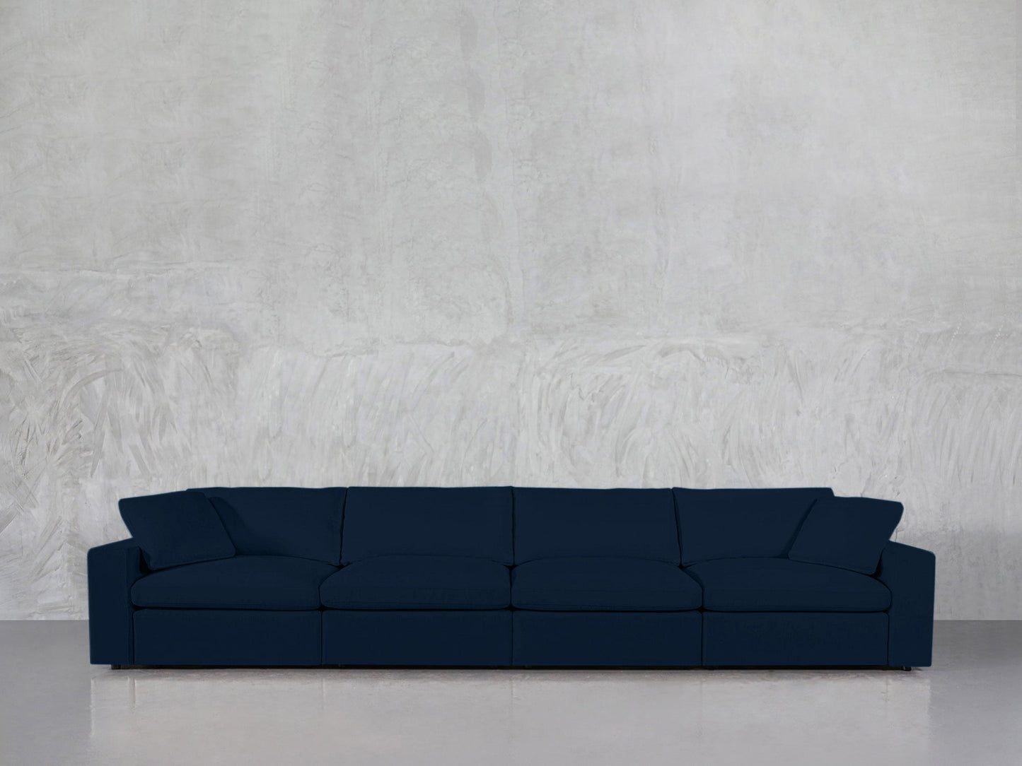 A 4-Seat Modular Sofa in dark blue with four seat cushions and two side pillows is placed against a light gray textured wall on a shiny floor.