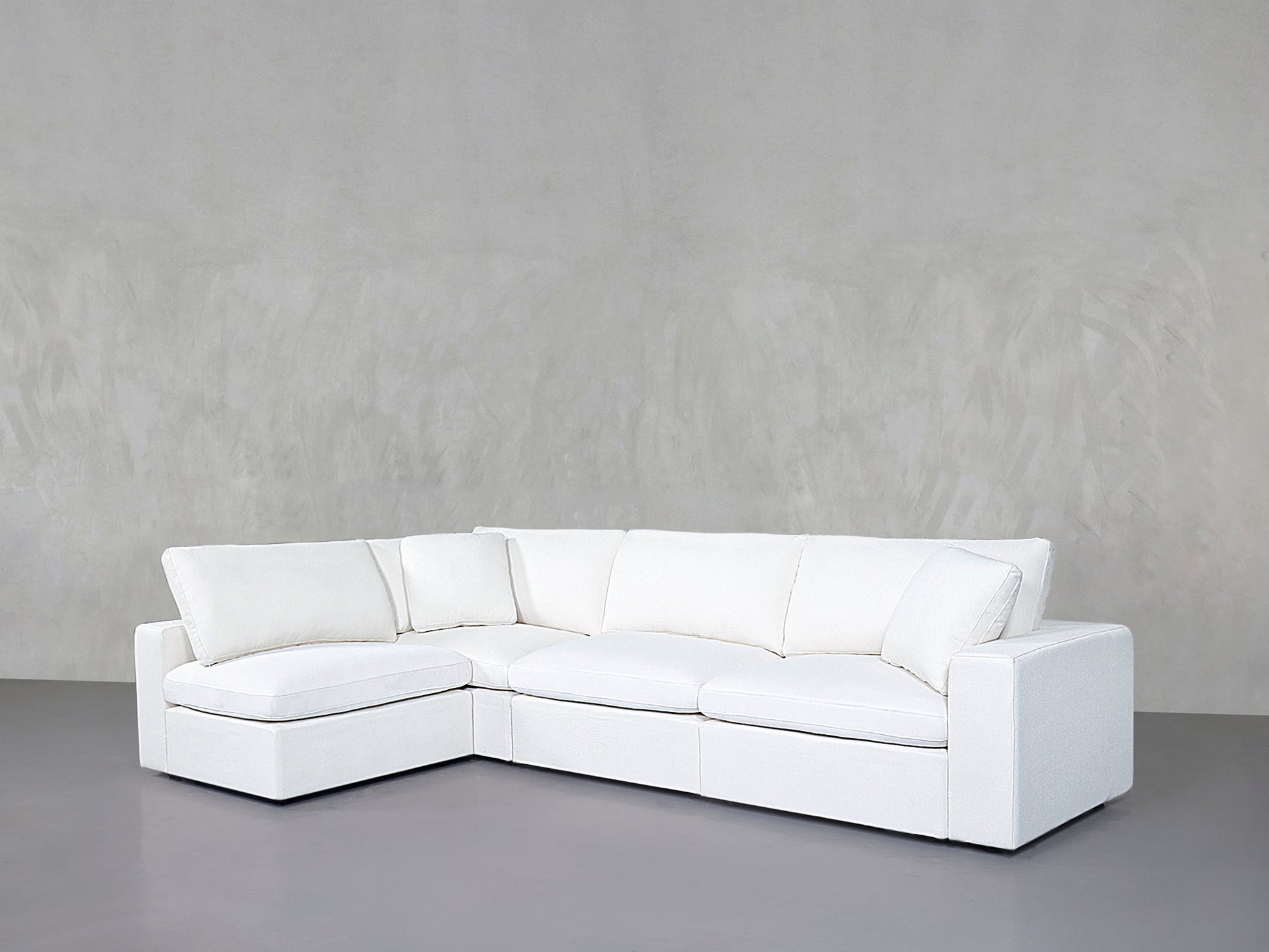 A 4-Seat Modular Open Corner Sectional in modern white with clean lines and large cushions, set against a plain light gray wall and gray floor.