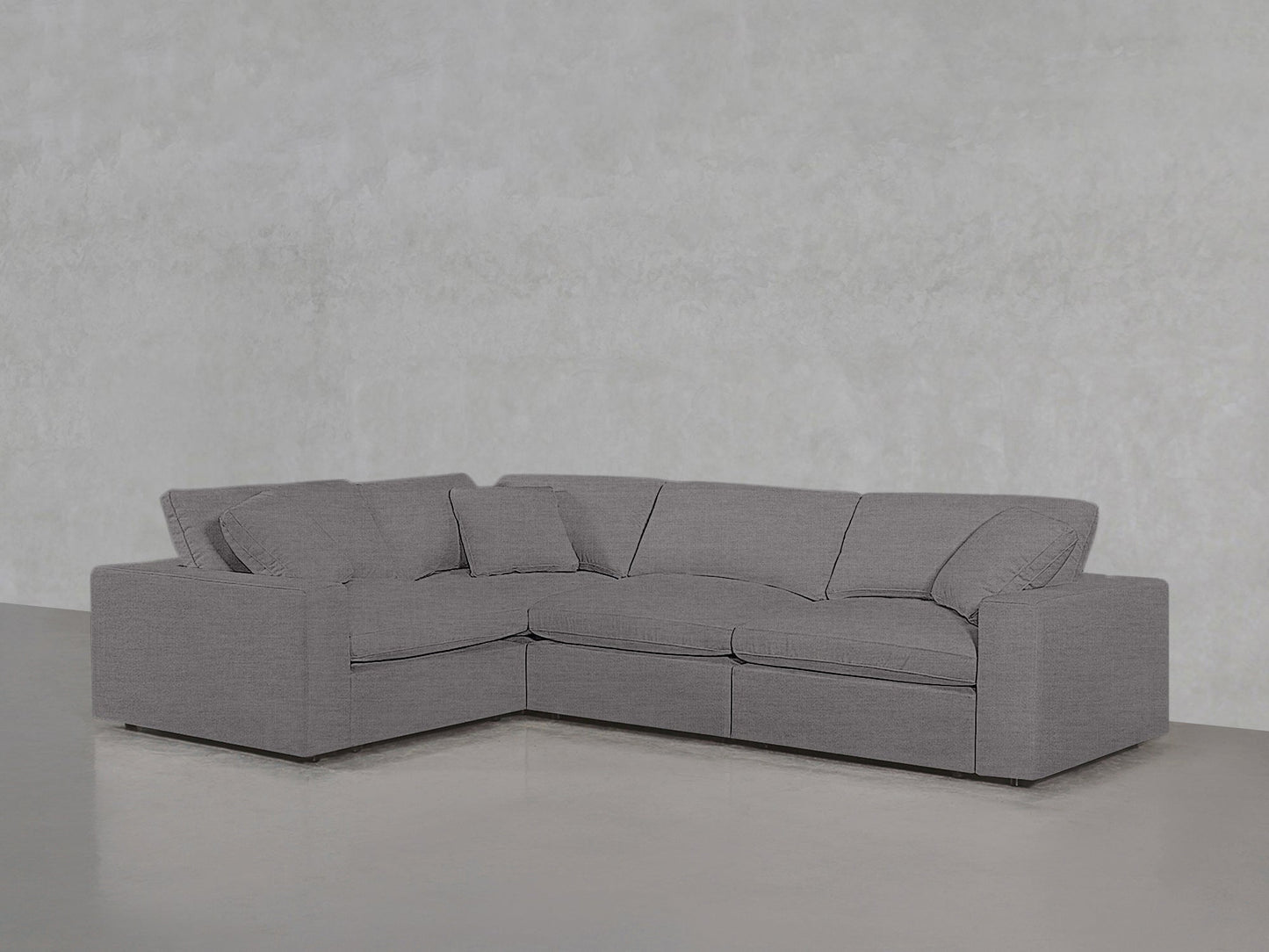 A 4-Seat Modular Corner Sectional with clean lines and plush cushions sits against a plain light gray textured wall on a smooth floor in a minimalist room.