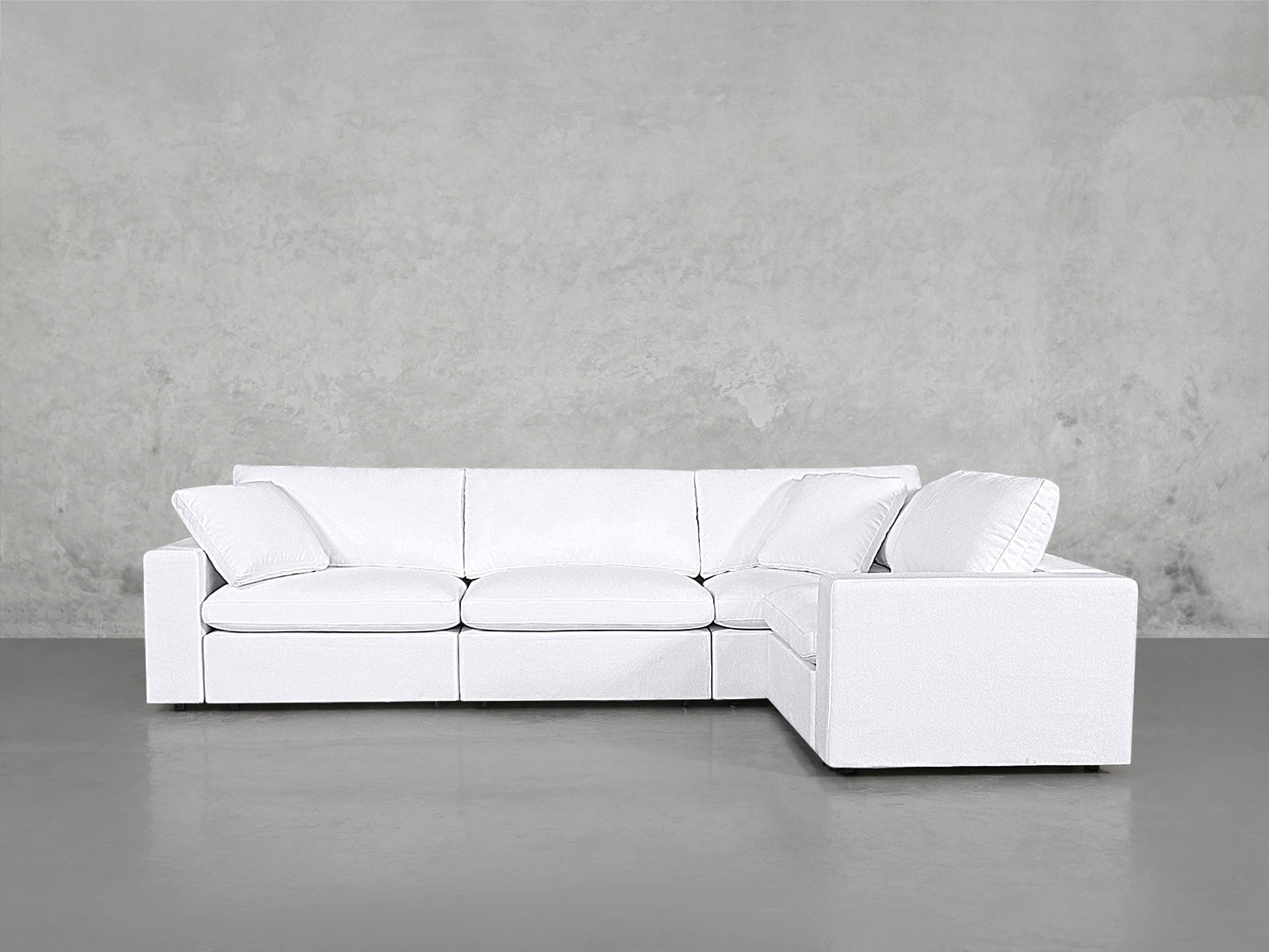 A 4-Seat Modular Corner Sectional in modern white with clean lines and plush cushions sits on a smooth gray floor against a light gray concrete wall.