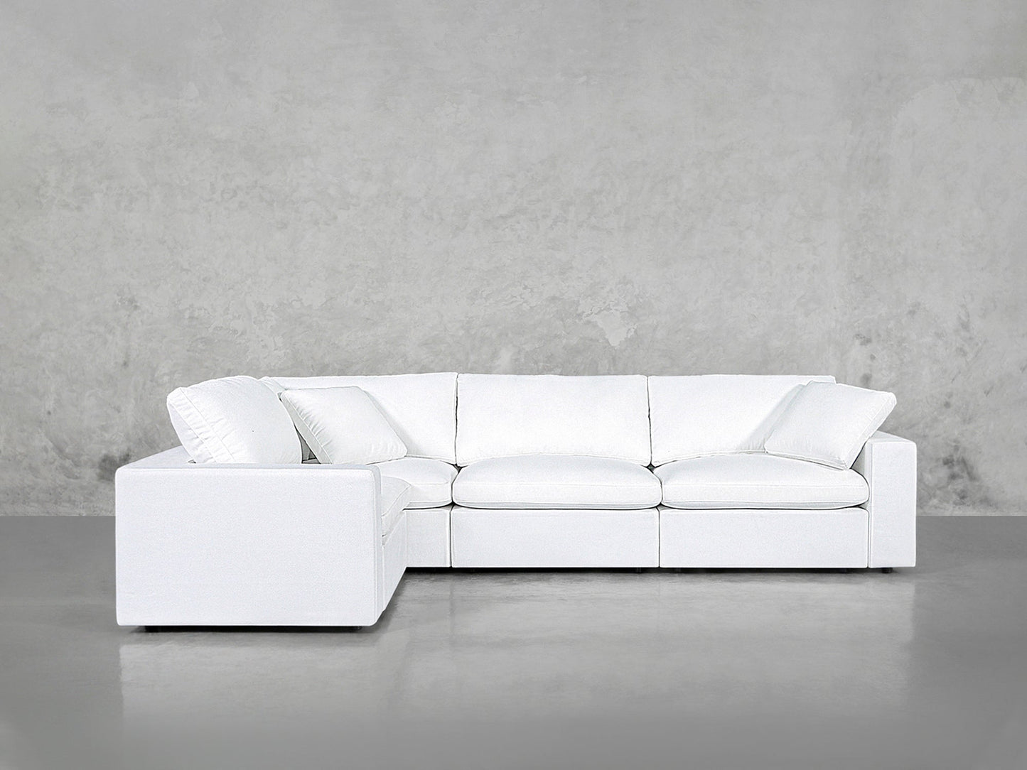 A 4-Seat Modular Corner Sectional with clean lines and a modern white finish is set against a plain, light gray concrete wall and floor.
