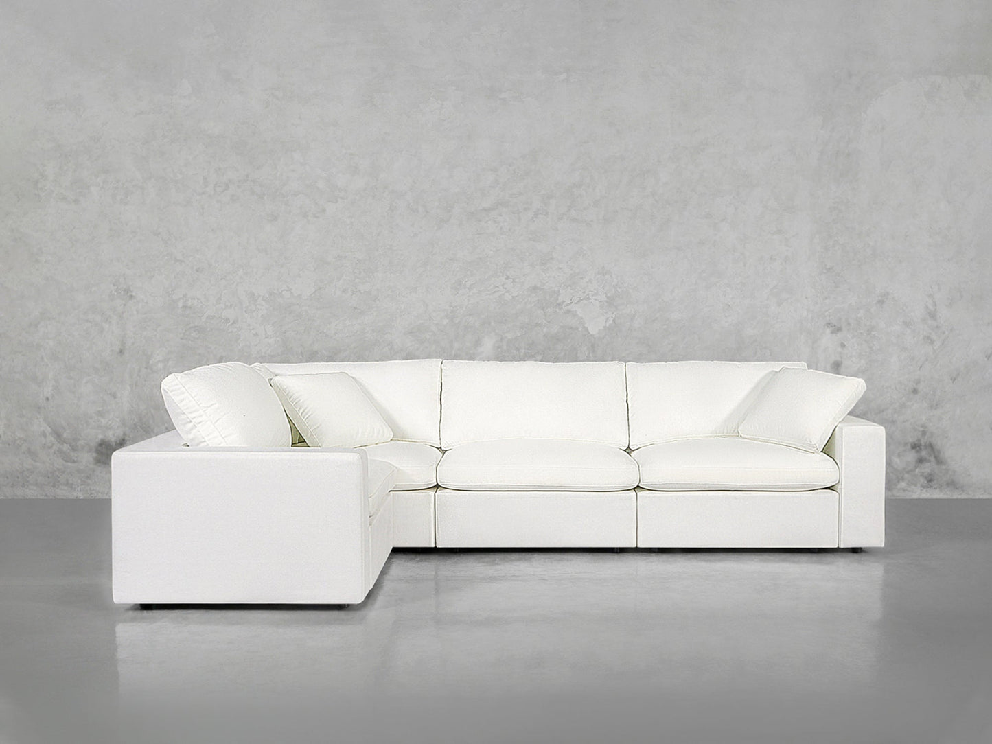 A modern 4-Seat Modular Corner Sectional in white, featuring three seat and back cushions, set against a plain light gray concrete wall and floor.