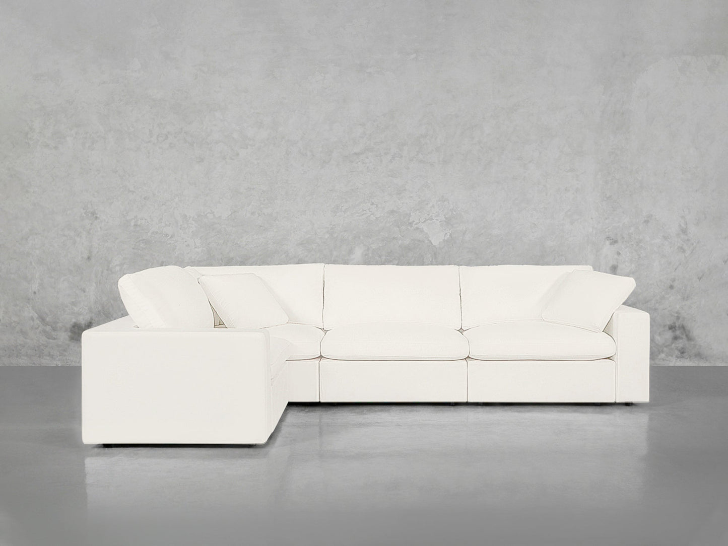 A 4-Seat Modular Corner Sectional with plush cushions and pillows stands in an empty room featuring a smooth gray concrete floor and a plain textured gray wall background.