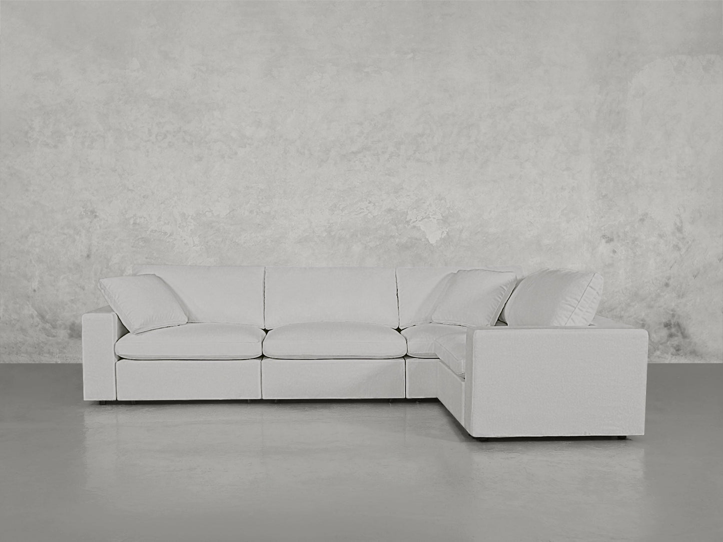 A 4-Seat Modular Corner Sectional in light gray with clean lines sits on a polished concrete floor against a textured, pale gray wall, featuring three seat cushions and an L-shaped design.