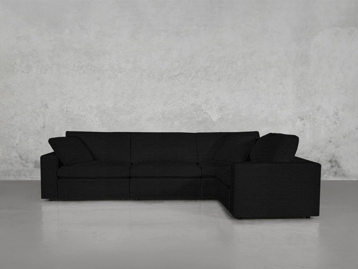 A 4-Seat Modular Corner Sectional in sleek black with clean lines and two back cushions, set against a light gray textured wall on a polished gray floor.
