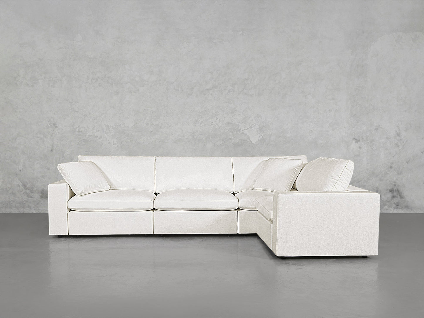A 4-Seat Modular Corner Sectional in white, featuring three seat cushions and two large back cushions, arranged in an L-shape against a plain concrete wall and grey floor in a minimalist space.