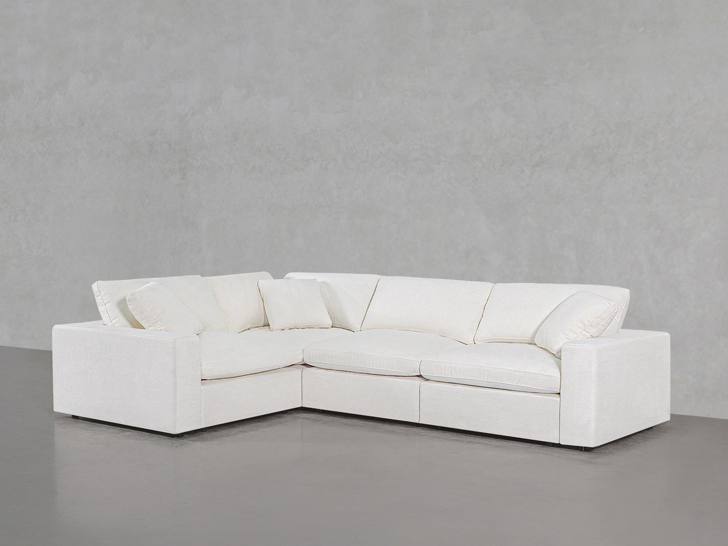 A 4-Seat Modular Corner Sectional with plush cushions and armrests, featuring an L-shaped white design, is set against a plain light gray wall on a smooth gray floor.