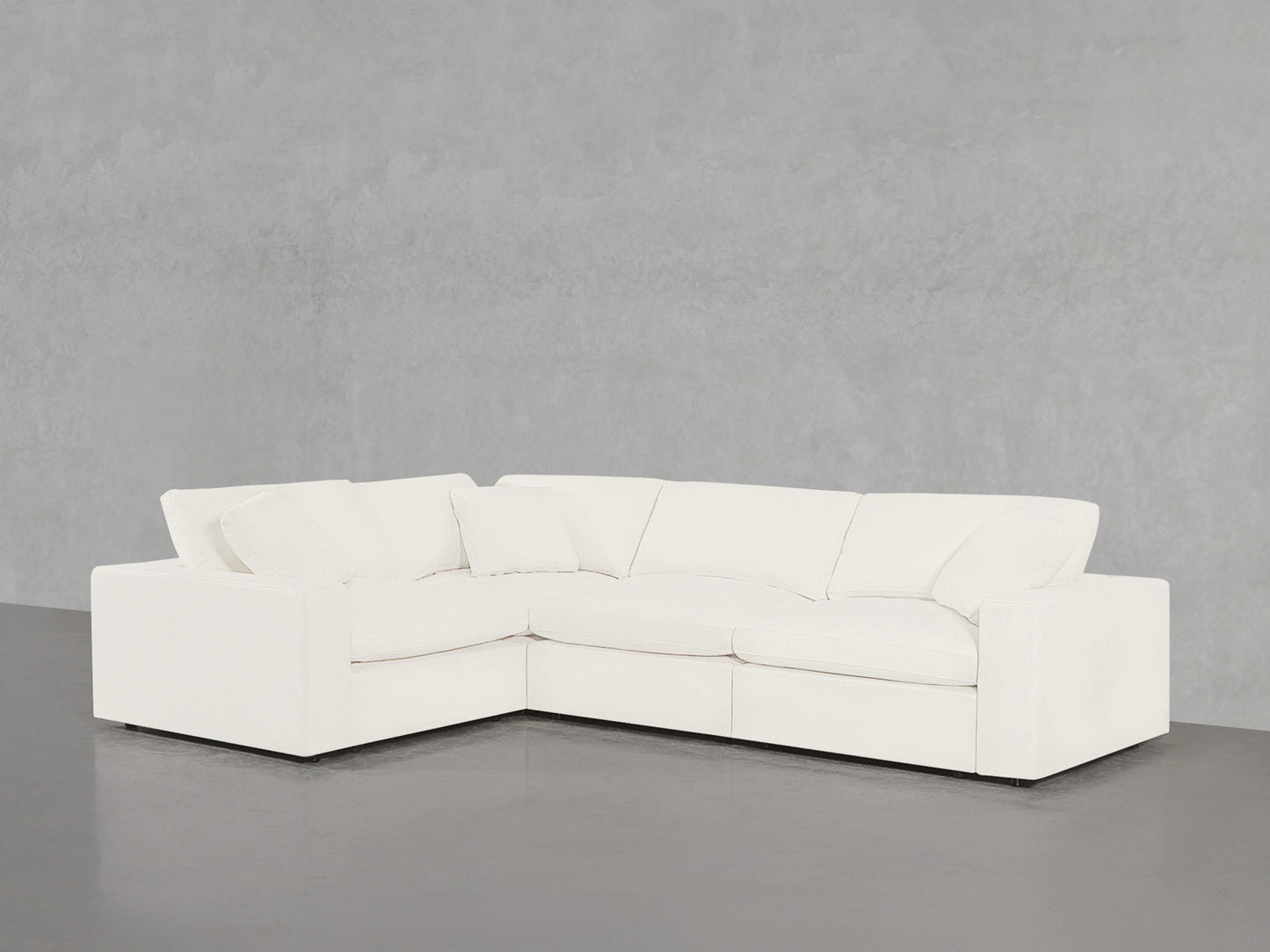A 4-Seat Modular Corner Sectional in white with three seat and three back cushions, set in a minimalist room featuring a plain gray wall and a dark gray floor.
