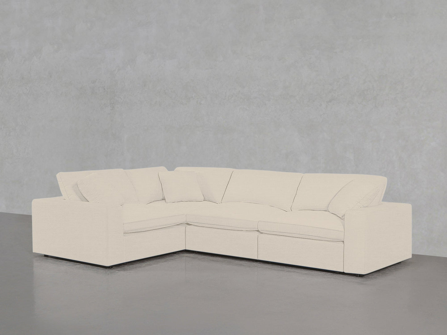 A 4-Seat Modular Corner Sectional with plush cushions and a cream finish sits on a smooth gray floor against a light gray textured wall.