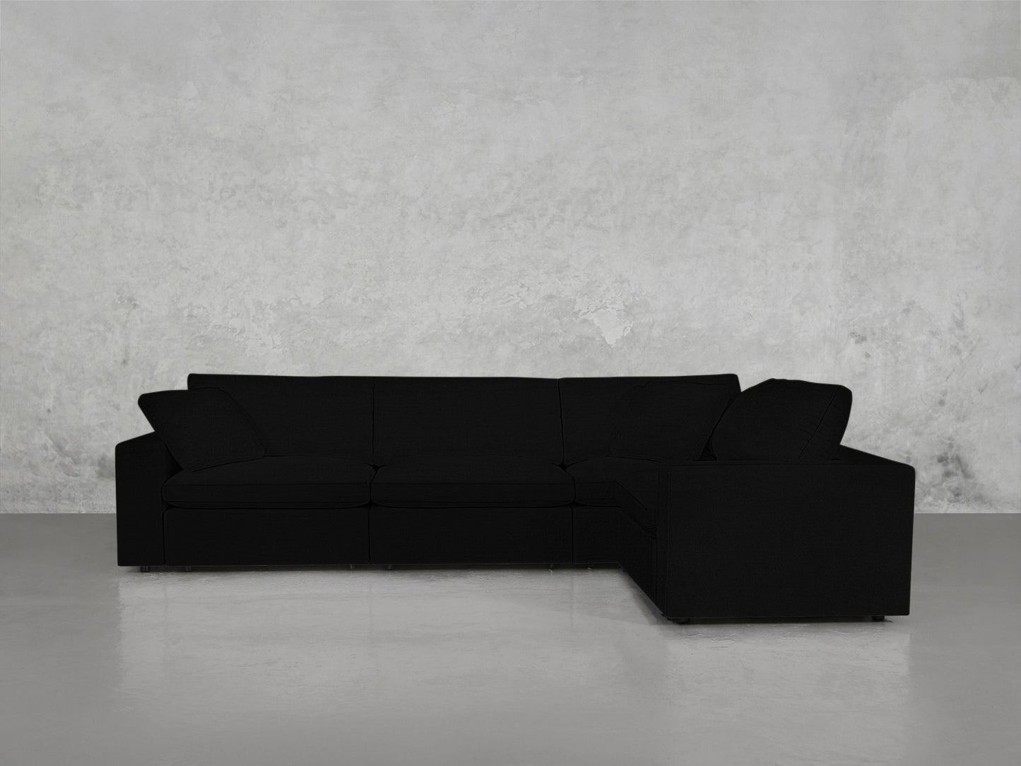 A 4-Seat Modular Corner Sectional with modern black upholstery and clean lines is set in a minimalist room featuring a polished concrete floor and a light gray textured wall.