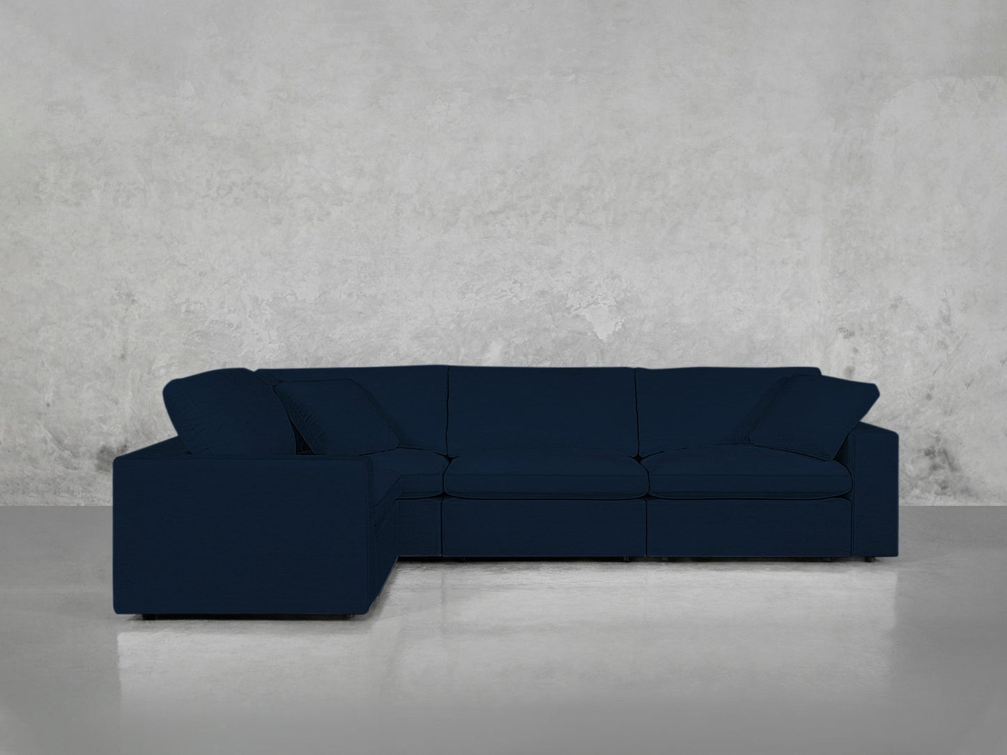 A 4-Seat Modular Corner Sectional in dark blue, complete with cushions, is set against a light gray textured wall and a polished, light-colored floor.