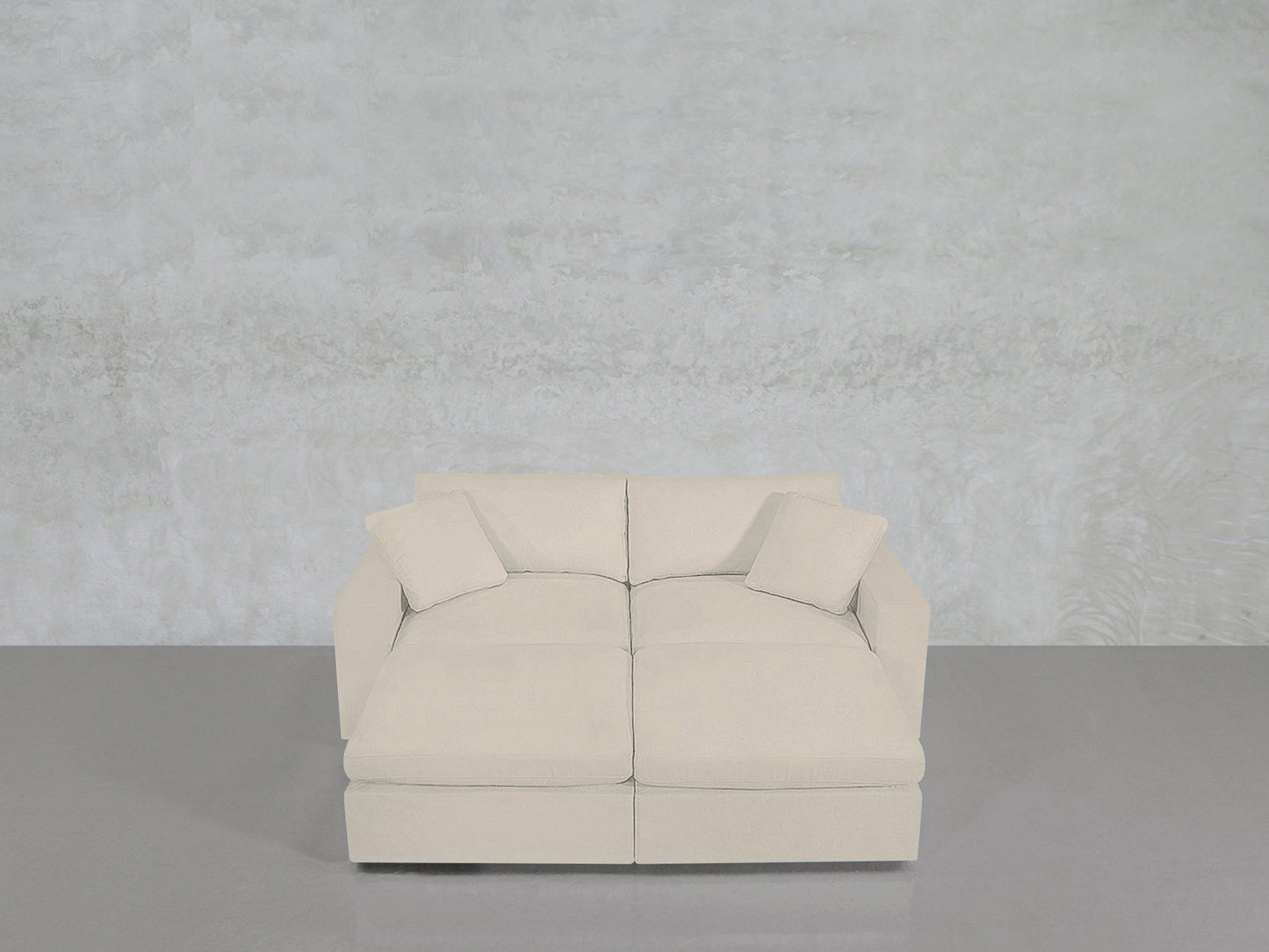 A light beige, modern 4-Seat Modular Daybed with two matching throw pillows sits on a smooth gray floor against a textured pale gray wall.