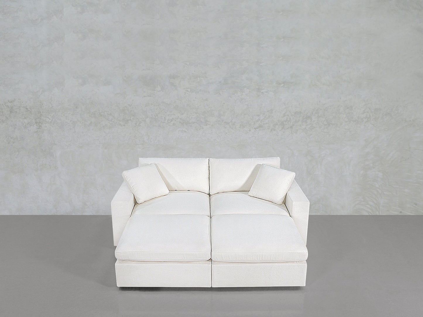 A modern, white 4-Seat Modular Daybed with two matching throw pillows sits on a smooth gray floor against a textured light gray wall.