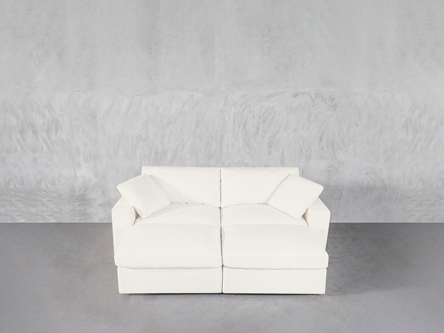 A modern white 4-Seat Modular Daybed with two seat cushions and two matching pillows sits against a textured light gray wall on a smooth gray floor.