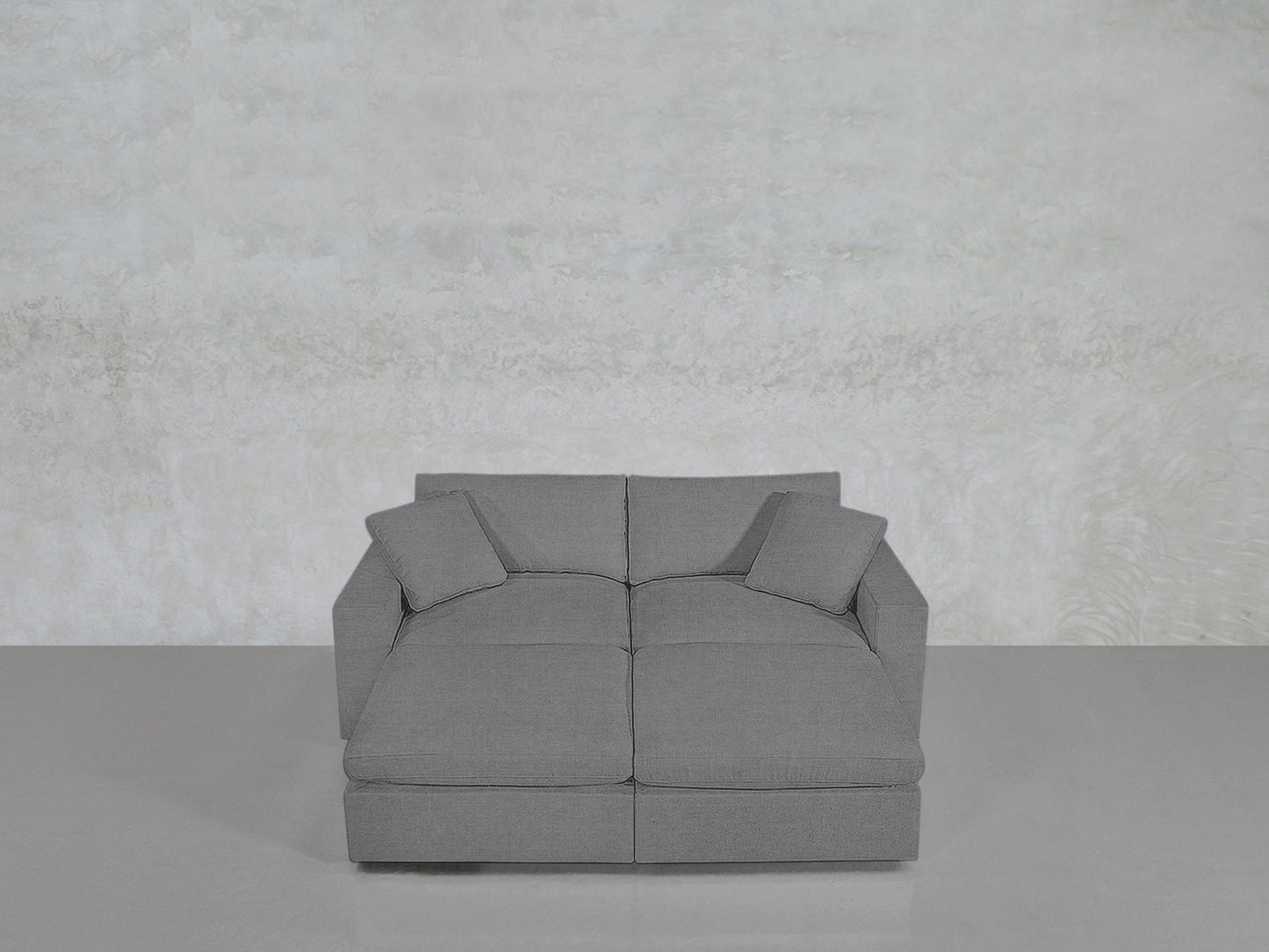 A 4-Seat Modular Daybed in modern gray with two matching cushions is set against a light textured wall and a smooth, shiny floor.