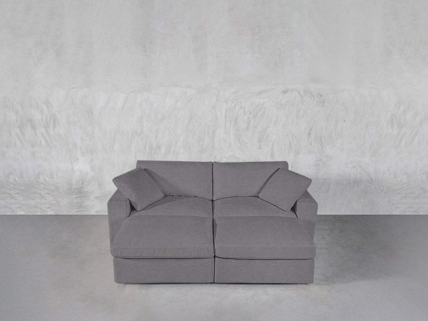 A 4-Seat Modular Daybed in modern gray with two seat cushions and two matching throw pillows is arranged against a textured light gray wall on a smooth gray floor.