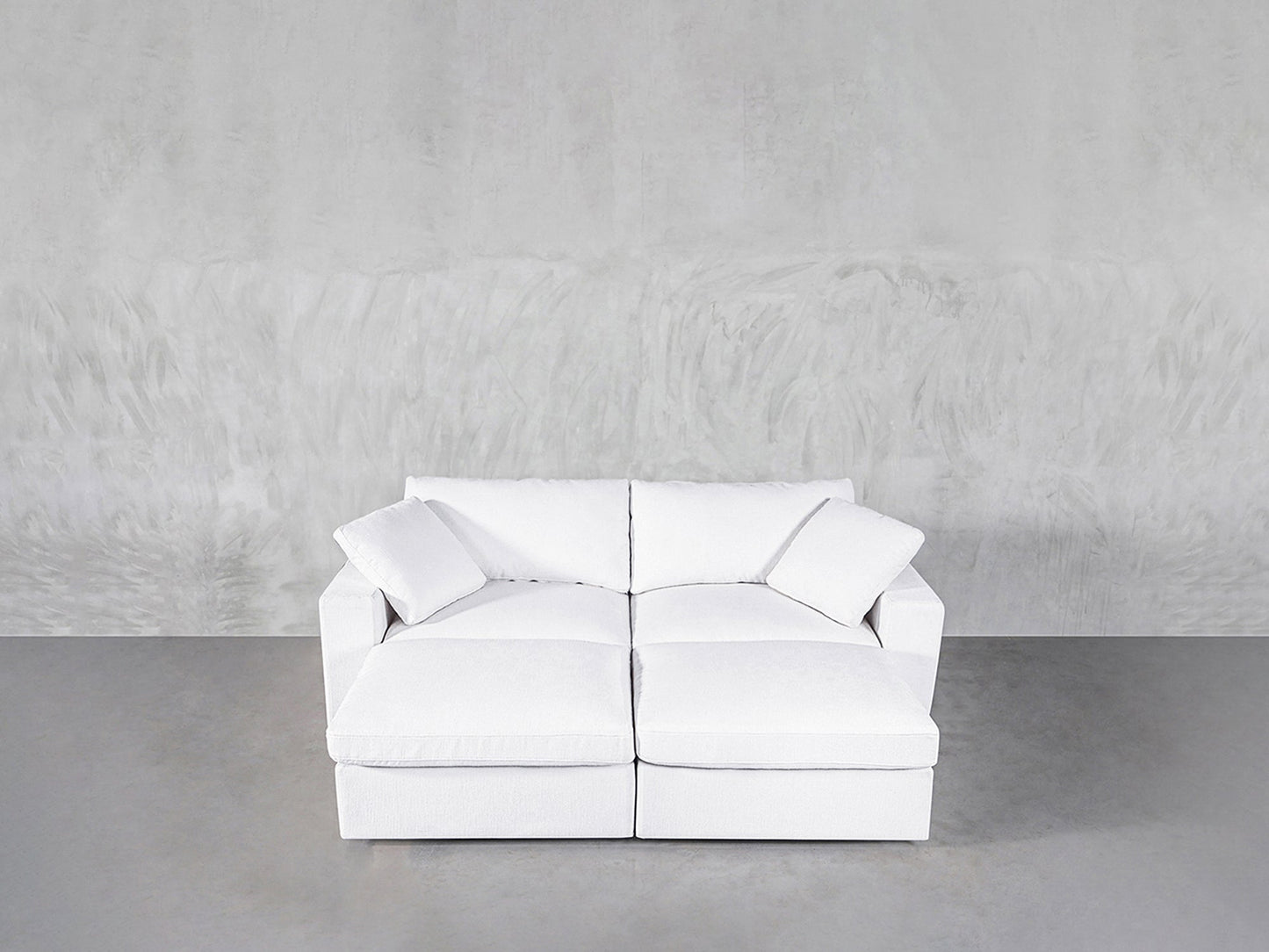 A modern white 4-Seat Modular Daybed with two seat and back cushions sits against a light gray textured wall on a smooth gray floor. Two matching white throw pillows are placed in each corner of the daybed.