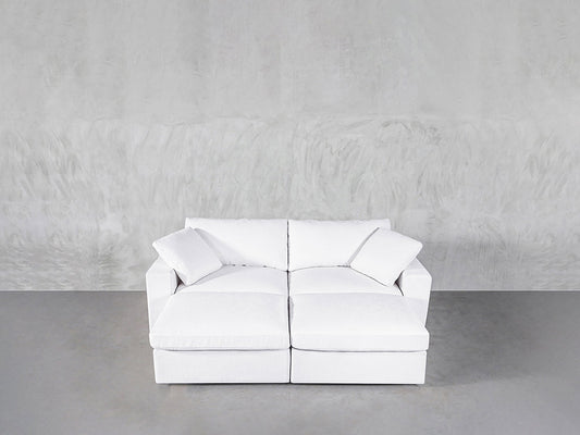 A modern white 4-Seat Modular Daybed with two seat and back cushions sits against a light gray textured wall on a smooth gray floor. Two matching white throw pillows are placed in each corner of the daybed.