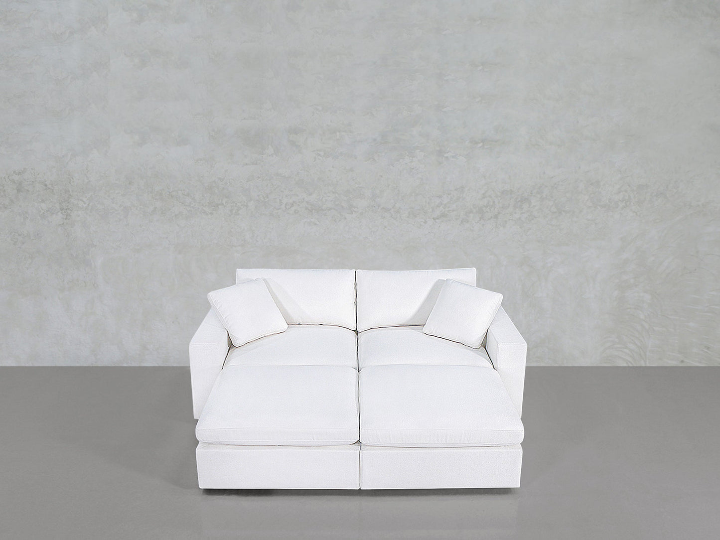 A modern, white 4-Seat Modular Daybed with two matching white cushions sits against a plain, light gray textured wall and a smooth gray floor.