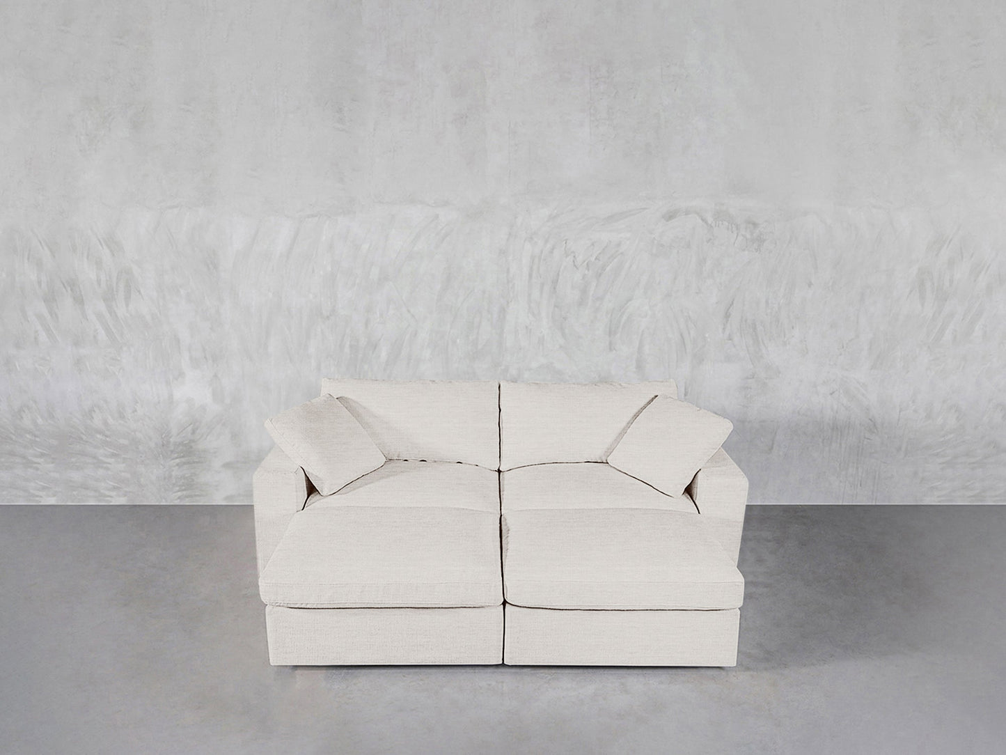 A 4-Seat Modular Daybed with an off-white finish, two seat cushions, and two matching throw pillows sits against a textured light gray wall on a polished concrete floor.