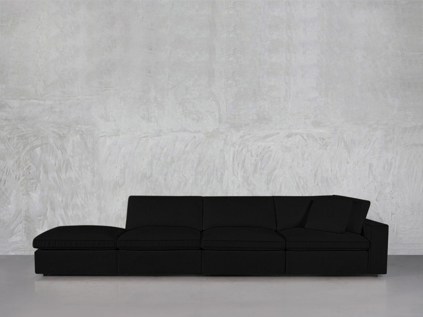 The 4-Seat Modular Lounger Sofa in black, featuring one armrest, sits against a light gray textured wall and rests on a smooth, light-colored floor.