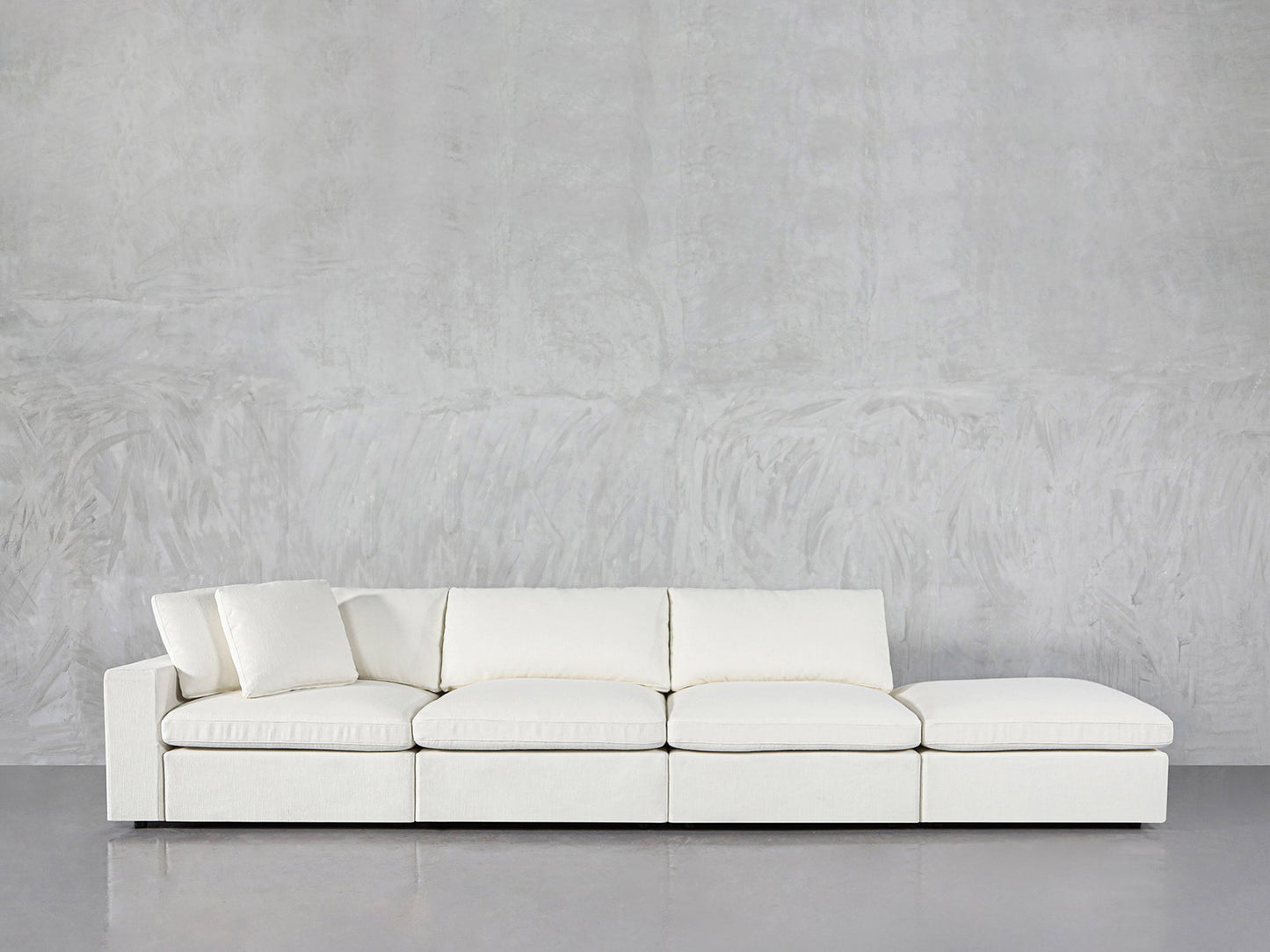 The 4-Seat Modular Lounger Sofa, modern and white with four sections and a matching cushion, sits against a textured light gray wall on a shiny gray floor.