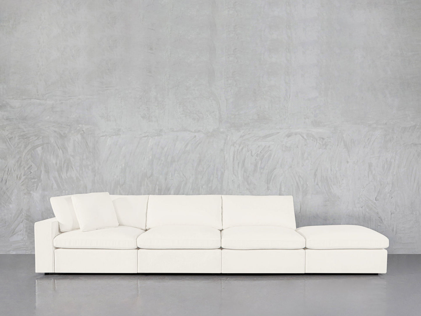 The 4-Seat Modular Lounger Sofa, featuring a modern white design with four seat sections and an angled back cushion, is placed against a textured light gray wall and a reflective gray floor.