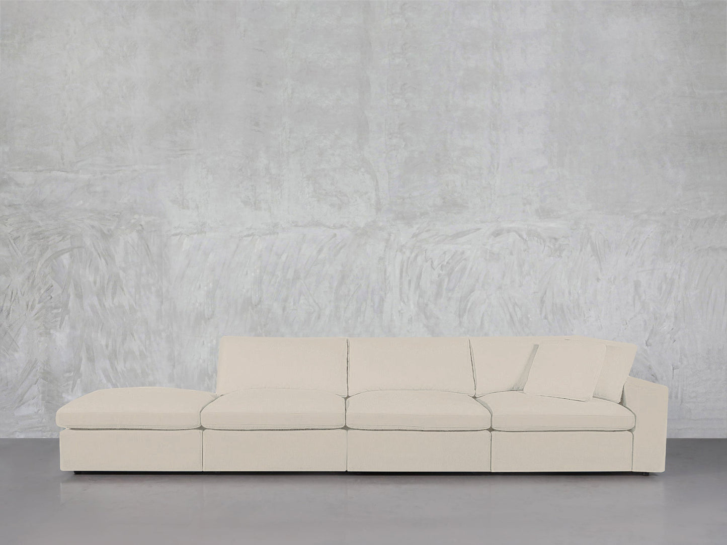 The 4-Seat Modular Lounger Sofa, in cream, with a matching cushion, is placed against a textured light gray wall on a polished concrete floor.