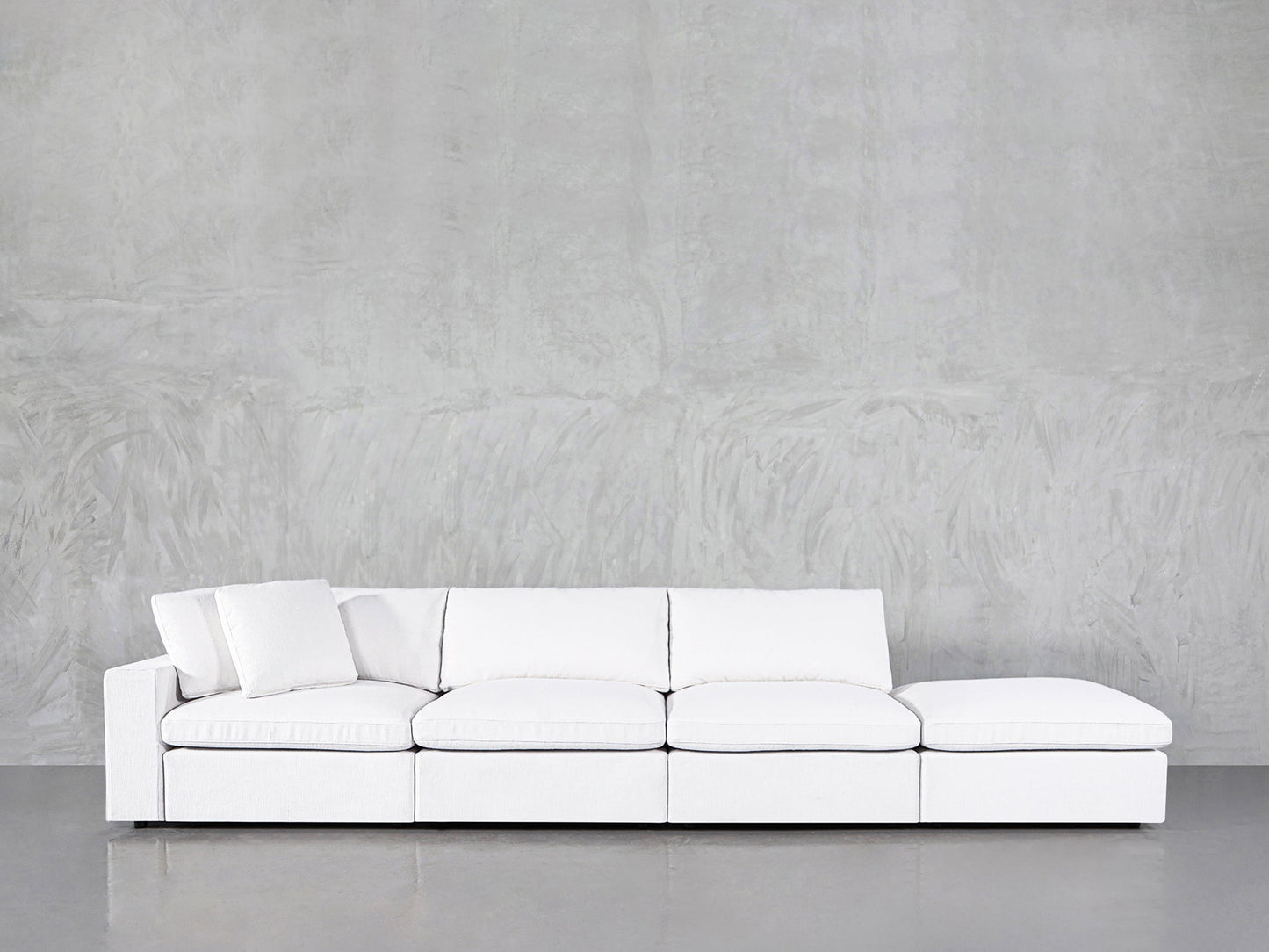The 4-Seat Modular Lounger Sofa features a modern white design with clean lines and three seat cushions, set against a light gray textured wall. A matching pillow sits on the sofa's left side.