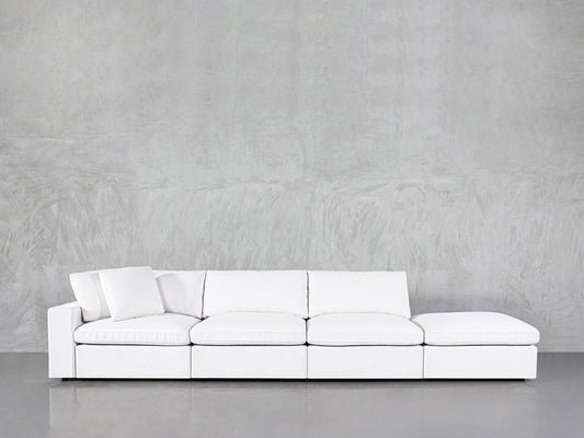 The 4-Seat Modular Lounger Sofa features a modern white design with clean lines and three seat cushions, set against a light gray textured wall. A matching pillow sits on the sofa's left side.