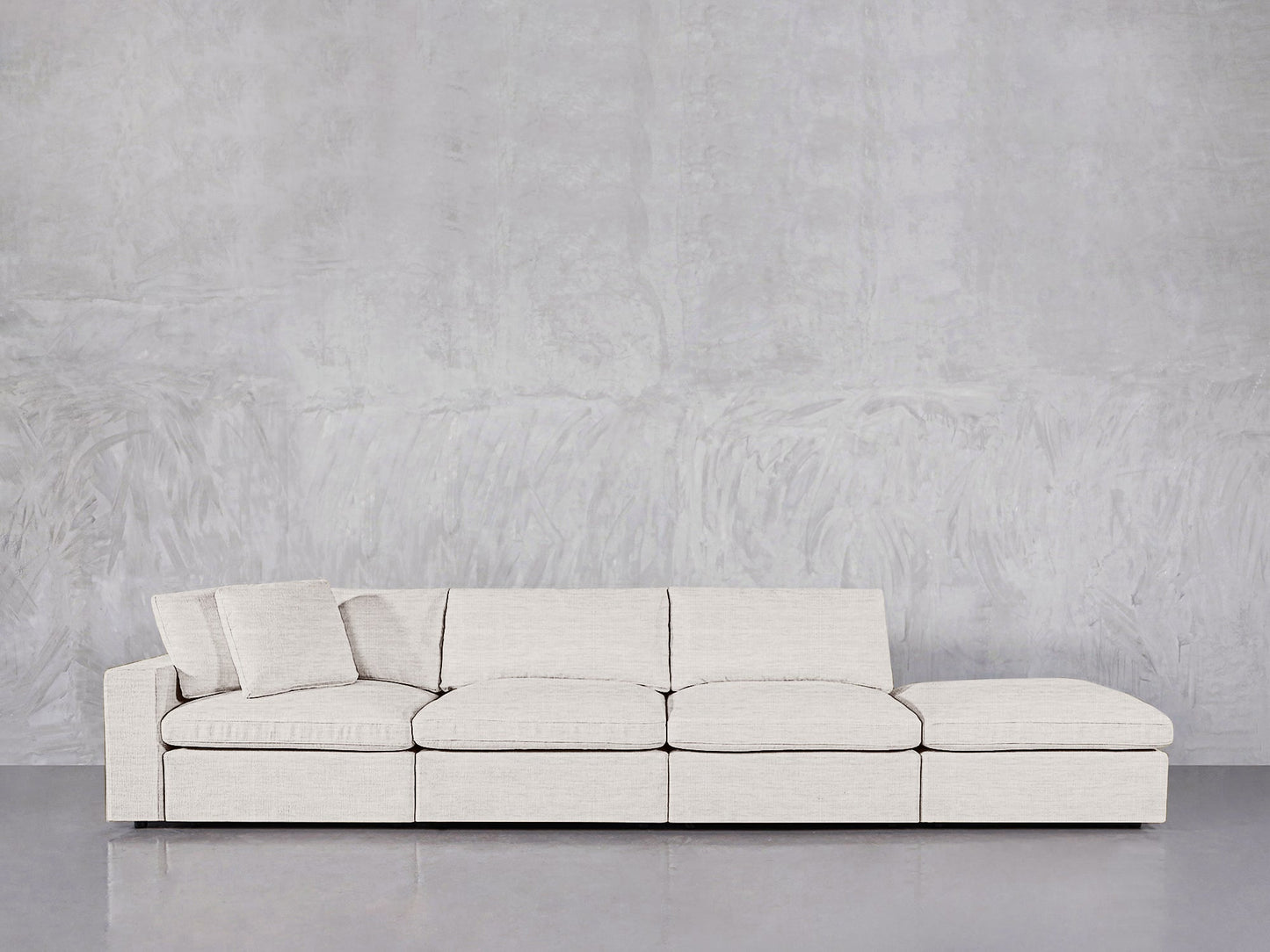 A modern 4-Seat Modular Lounger Sofa in light beige, featuring a chaise lounge and angled backrest pillow, sits against a plain textured gray wall on a reflective floor.