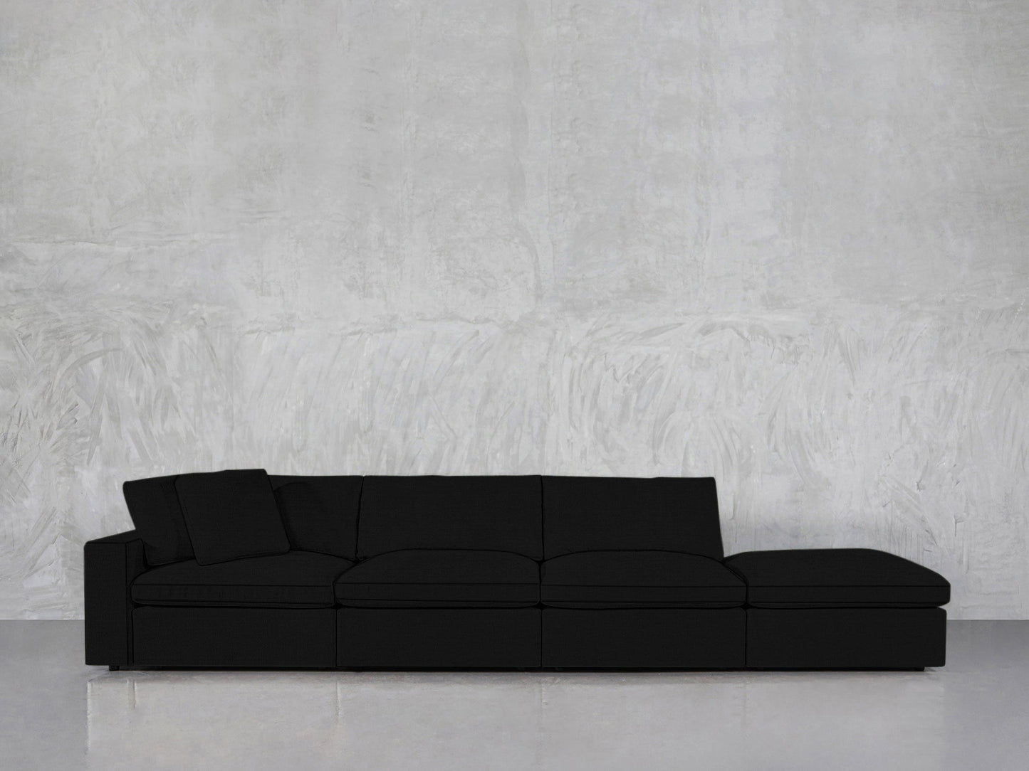 A 4-Seat Modular Lounger Sofa in black with clean lines and a matching cushion sits against a textured light gray wall on a polished floor.